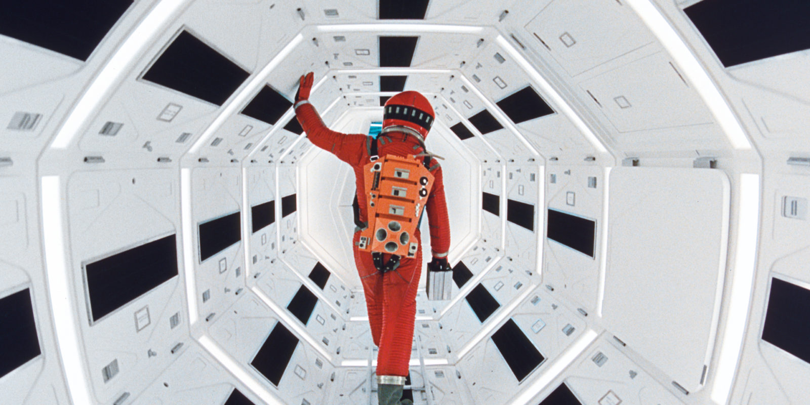 Die Rückenansicht eine Person in einem orangenen Astronautenanzug in einem achteckigen Gang in schwarz und weiß.
