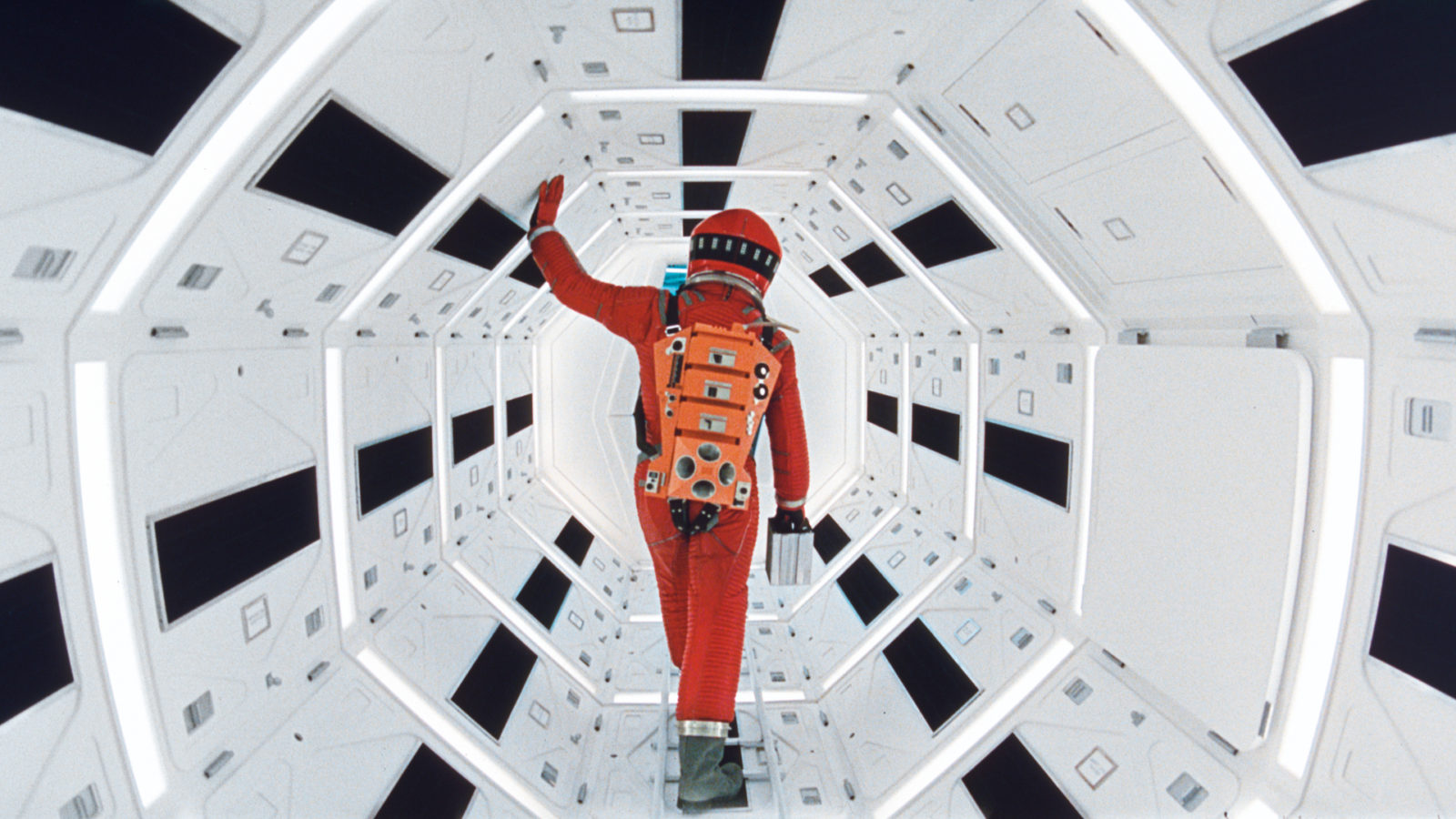 Die Rückenansicht eine Person in einem orangenen Astronautenanzug in einem achteckigen Gang in schwarz und weiß.