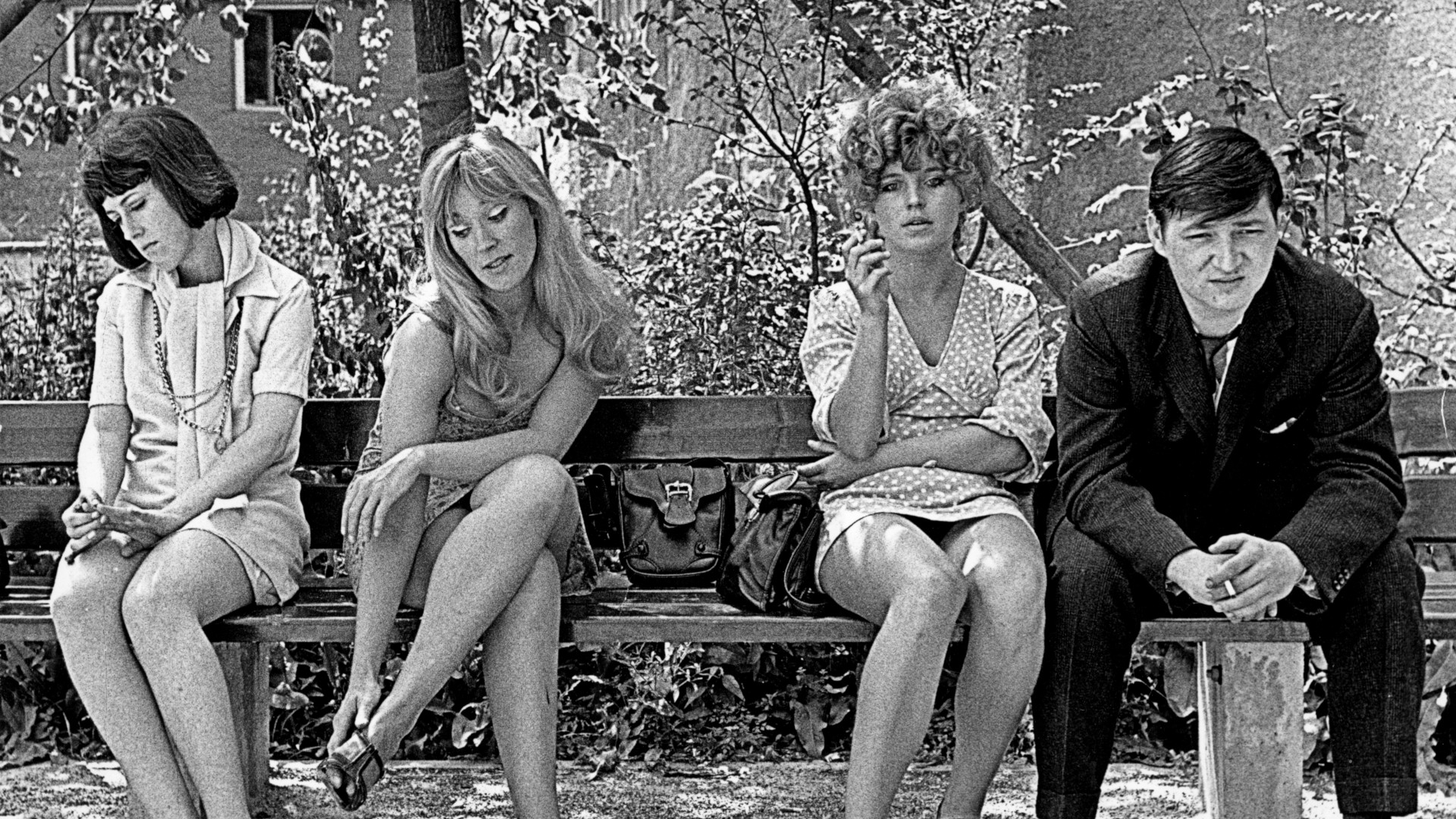 Filmstill aus dem Schwarz-Weiß-Film Katzelmacher. Drei Frauen sitzen auf einer Bank zusammen mit einem schwarz gekleideten Mann.