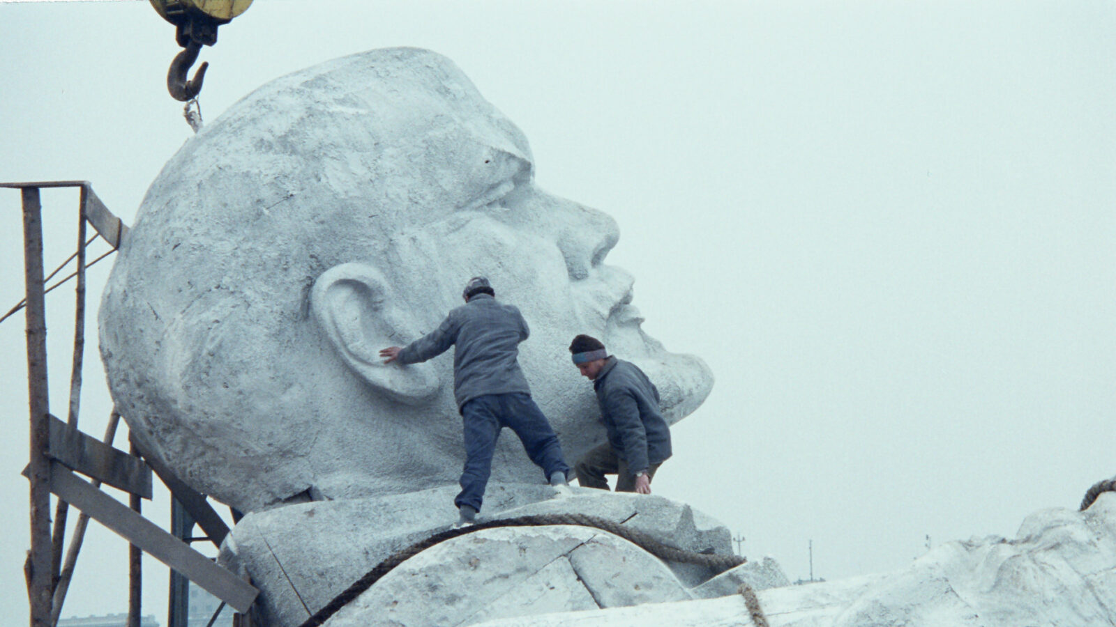 Filmstill aus den Film Symposium. Zwei Männer befinden sich auf dem Kragen einer Statur, die an einem Kran hängt. Die Statur stellt den russischen Politiker und kommunistischen Revolutionär Lenin sah.