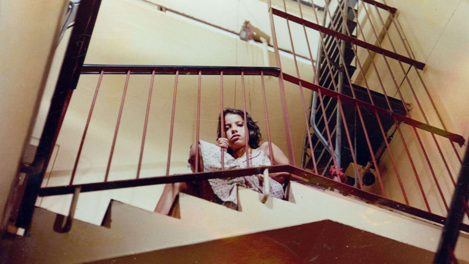 Filmstill aus Isabel auf der Treppe. Ein Mädchen sitzt hinter dem Treppengeländer und schaut nach unten.