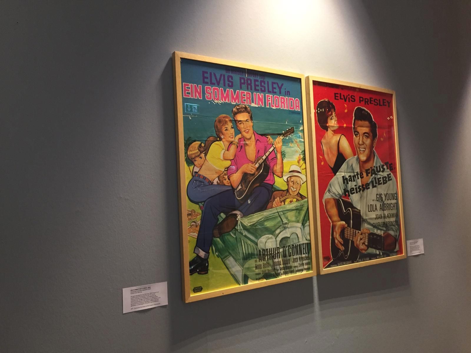 Blick in die Elvis-Ausstellung. Nebeneinander hängen zwei gelbe Bilderrahmen an einer grauen Wand. Beide zeigen Plakate zu Elvis.