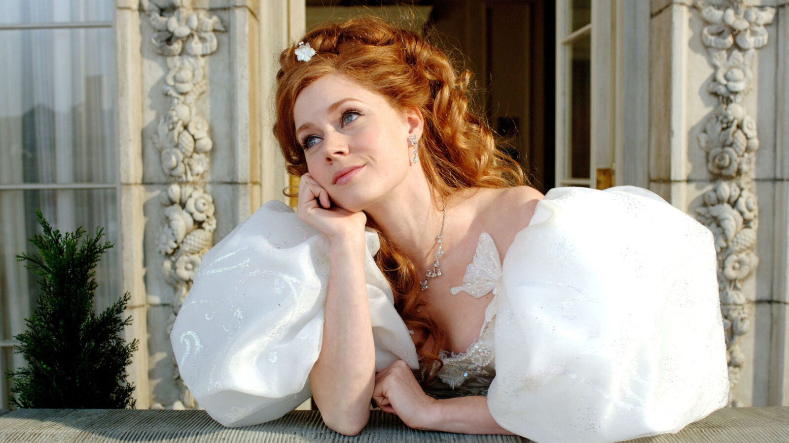 Filmstill aus Enchanted. Eine junge Frau in einem weißen pompösen Kleid steht auf einer Terrasse und stützt die Ellbogen auf die abgrenzende Mauer.