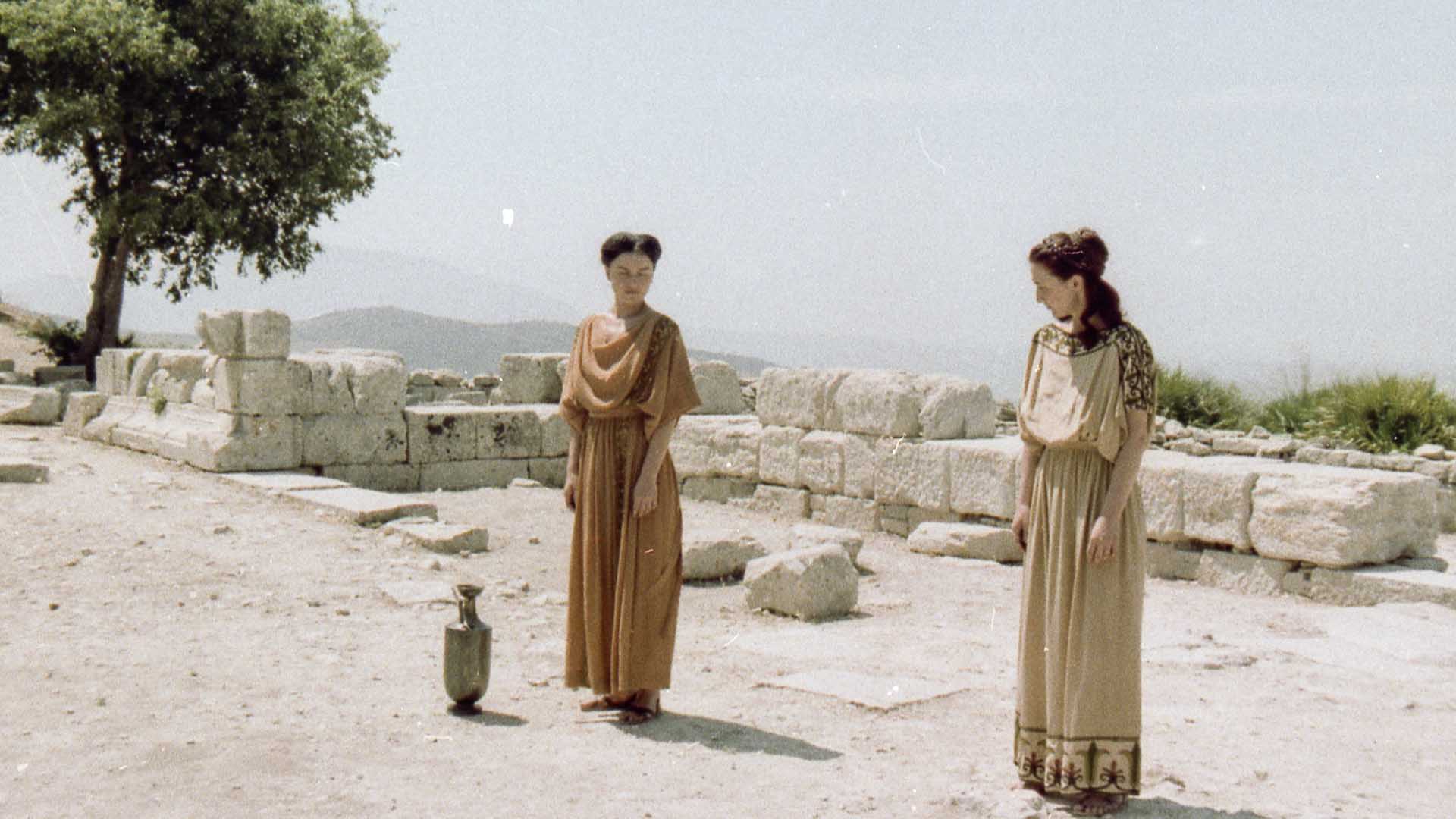 Filmstill aus Antigone. Zwei Frauen stehen in beigen Kleidern nebeneinander und schauen sich an. Die Umgebung zeigt eine niedrige Steinmauer, einen Baum und sandigen Boden.