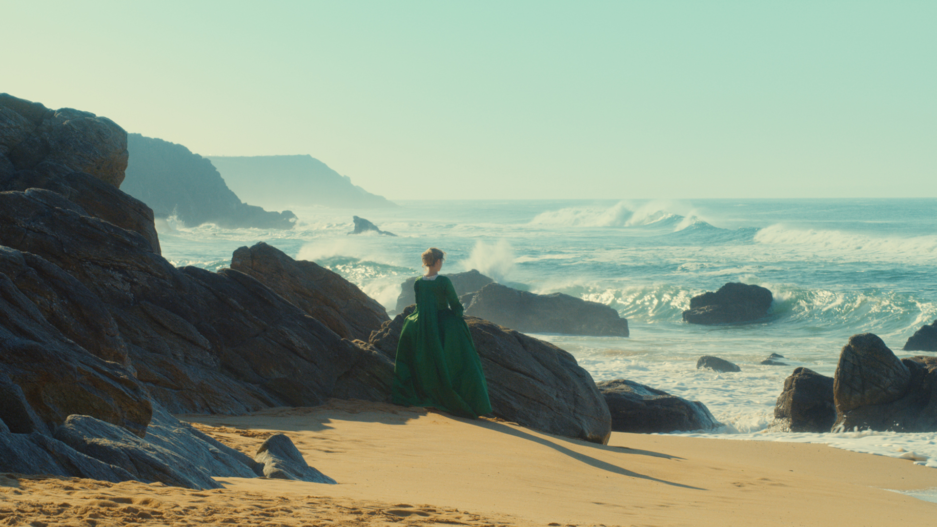 Filmstill Portrait einer jungen Frau in Flammen. Eine felsige Strandlandschaft und das Meer sind zu sehen. Eine junge Frau in grünem Kleid steht zwischen den Felsen.