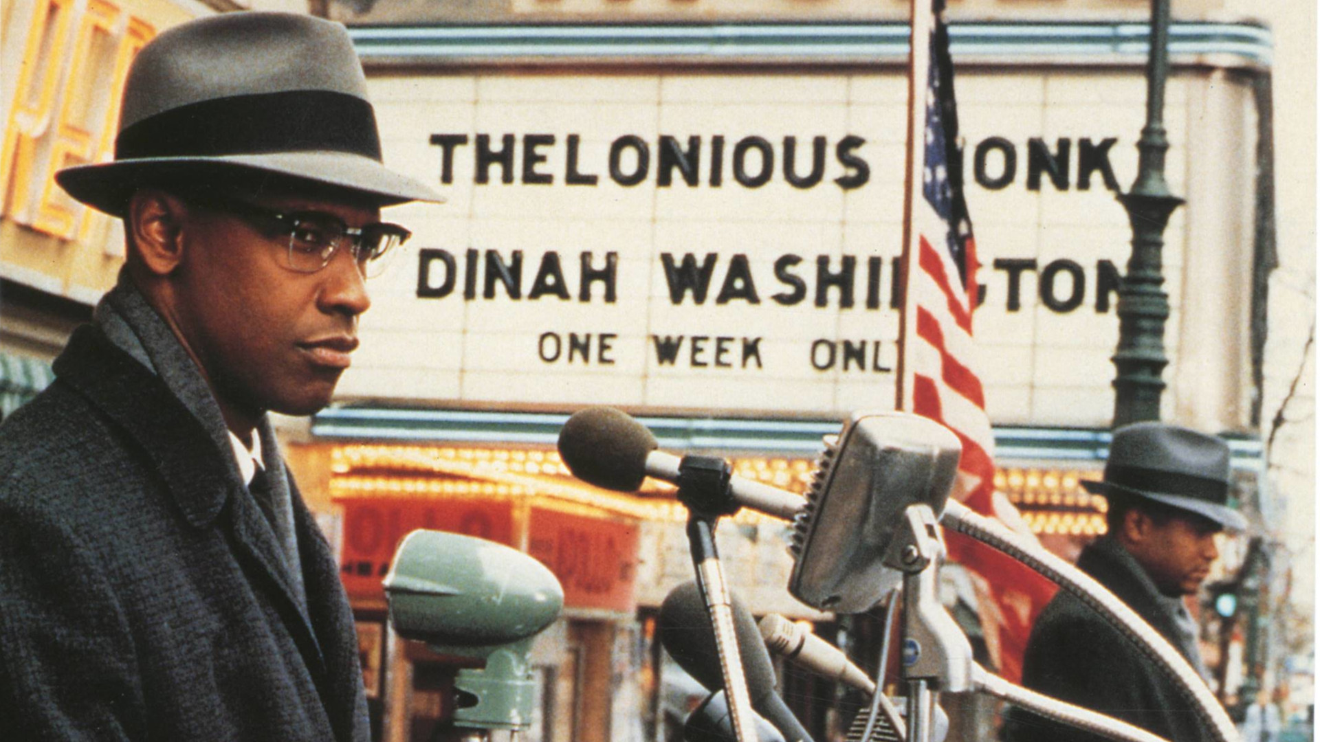 Filmstill aus Malcolm X. Ein Mann steht in Hut und Mantel hinter mehreren Mikrophonen und im Hintergrund sind die Flagge der USA und ein Werbelogo zu sehen.