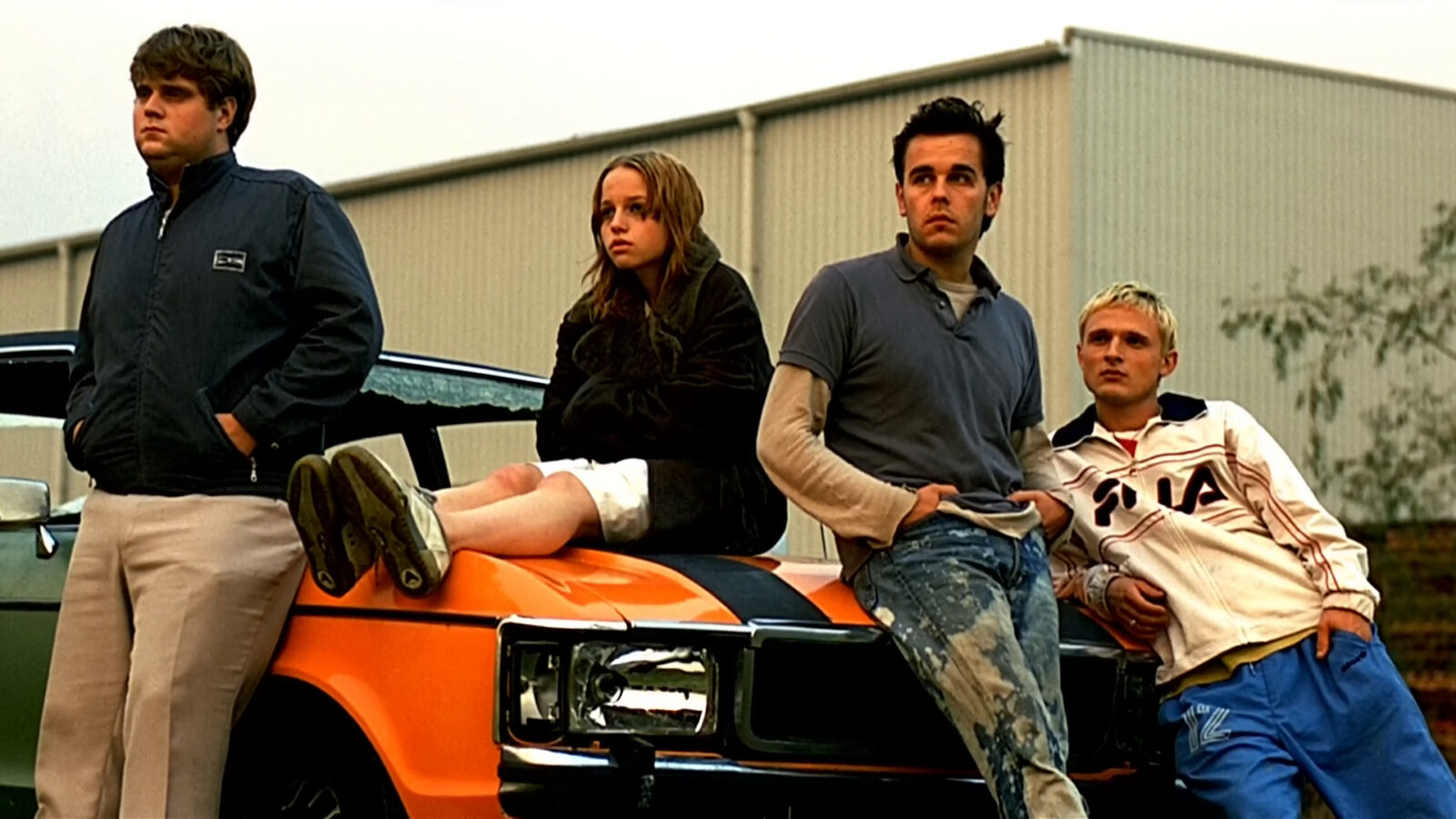 Filmstill aus Absolute Giganten. An einem orangenen Auto lehnen drei junge Männer. Auf der Motorhaube sitzt ein Mädchen.