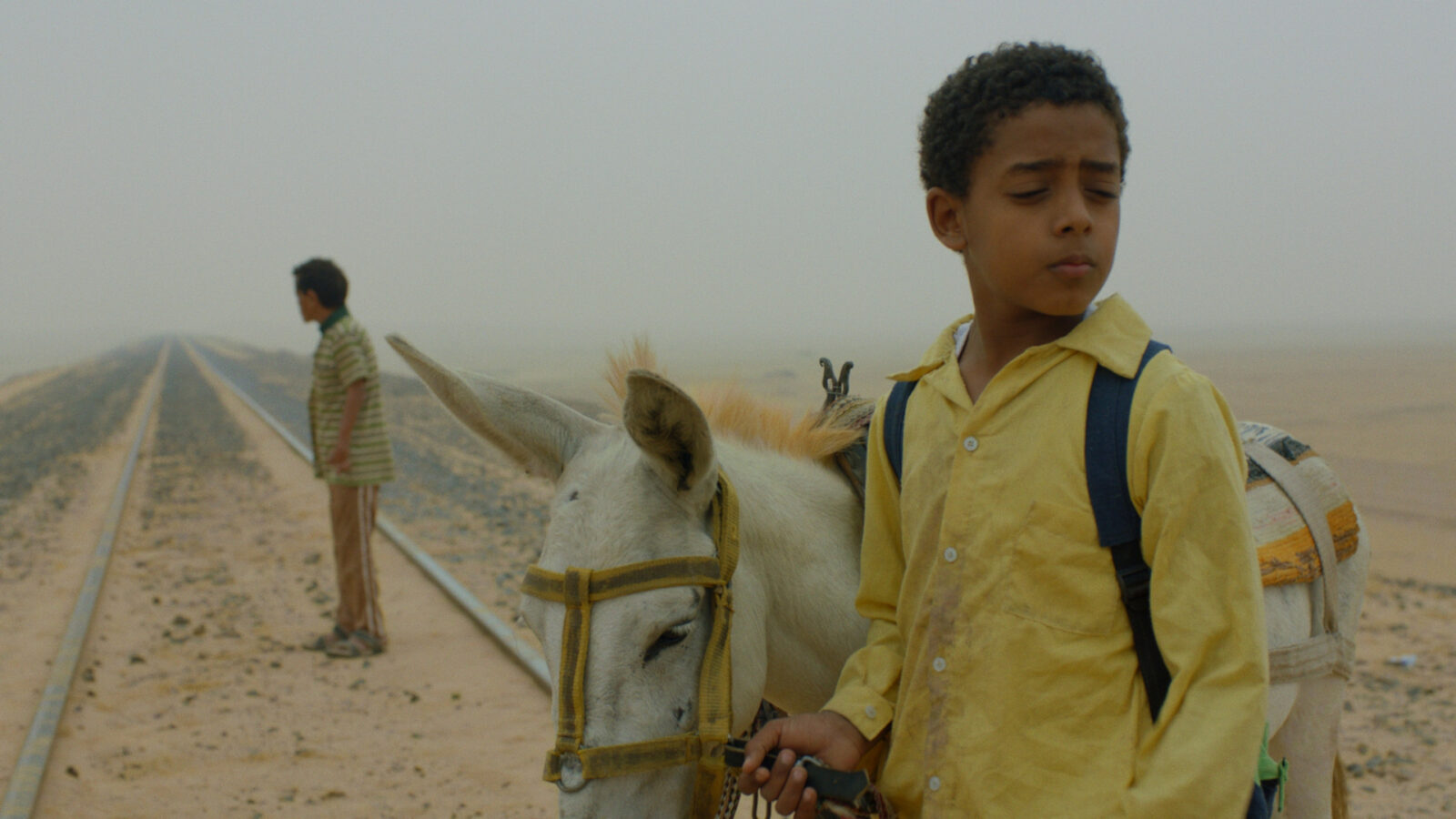 Filmstill aus dem Film Yomeddine. Im Vordergrund steht ein Junge mit einem Esel an der Hand auf einer Bahnschiene. Im Hintergrund steht eine Mann ebenfalls auf der Bahnschine, auf der viel Sand liegt.