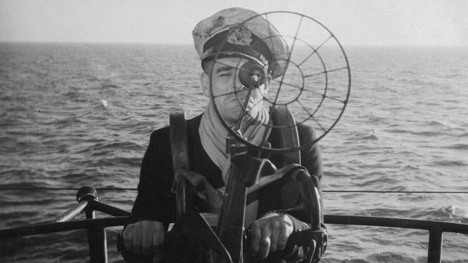 Filmstill Das Boot der Verdammten. Ein Mann mit Kapitänsmütze steht a ei e Gerät, durch das er hindurch schaut. Im Hintergrund befindet sich nur Wasser bis zum Horizont.