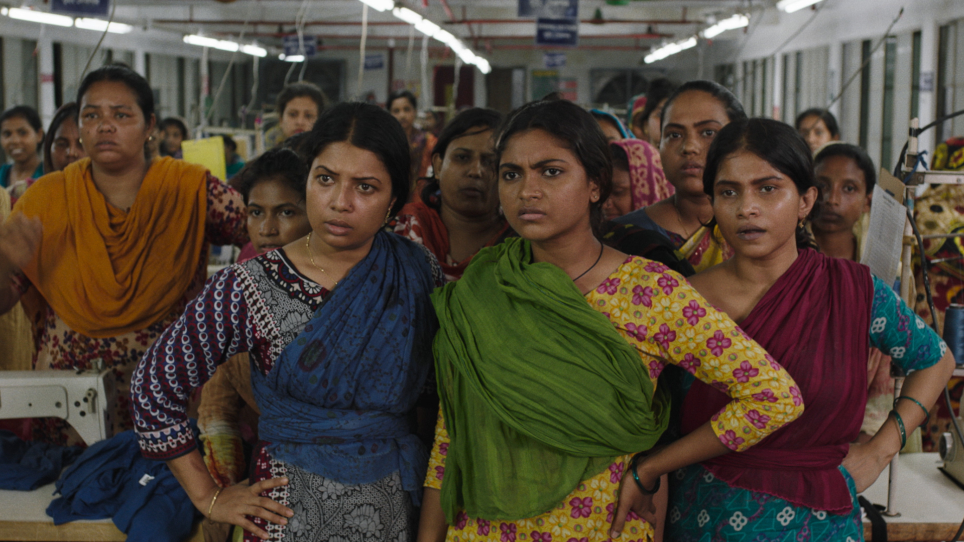 Filmstill Made in Bangladesh. Mehrere Frauen stehen vor der Kamera. Sie sind in bunte Klamotten gekleidet.