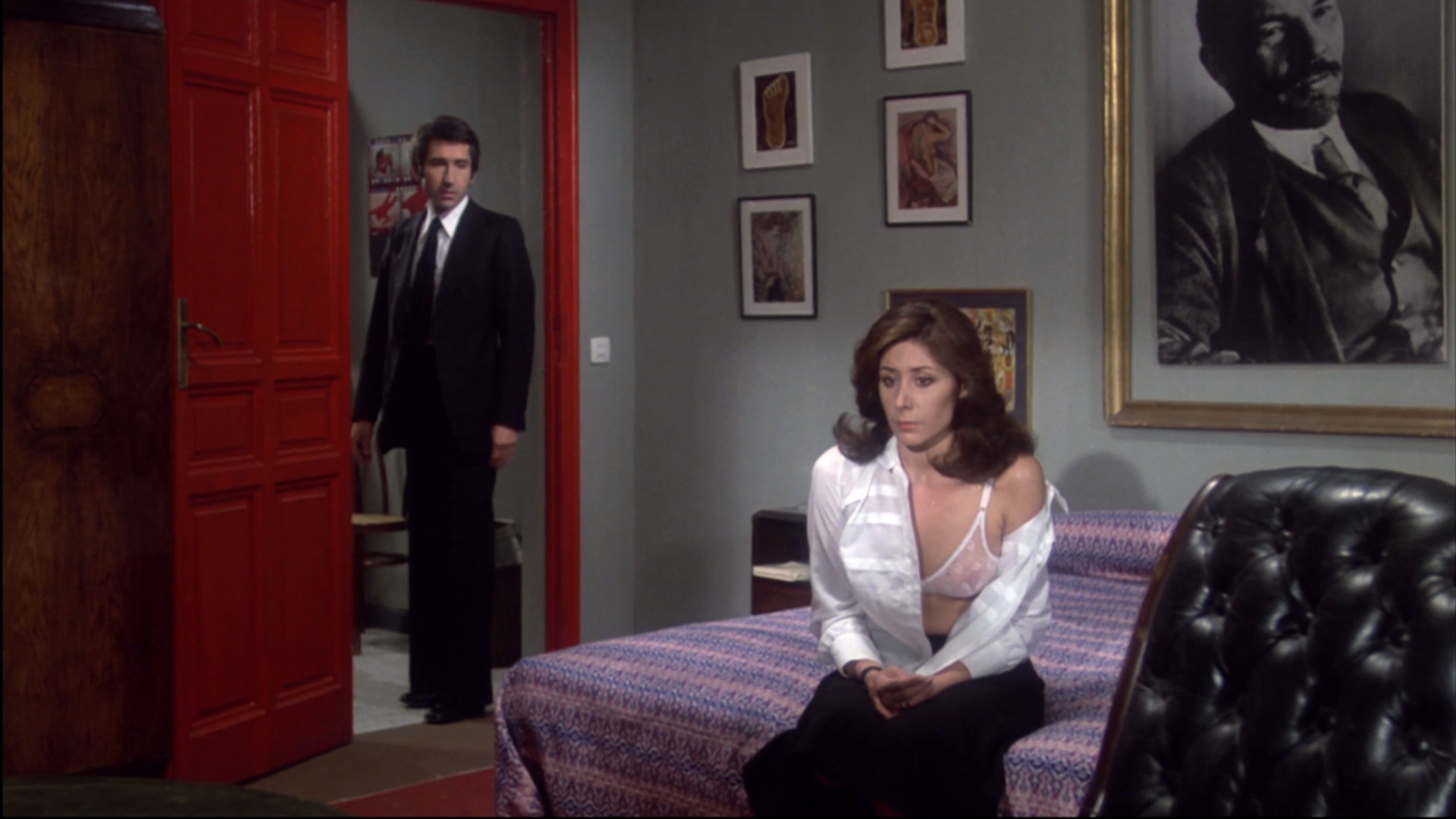 Eine Frau sitzt auf einem Bett und starrt vor sich hin. In der roten Zimmertür steht ein Mann im Anzug.