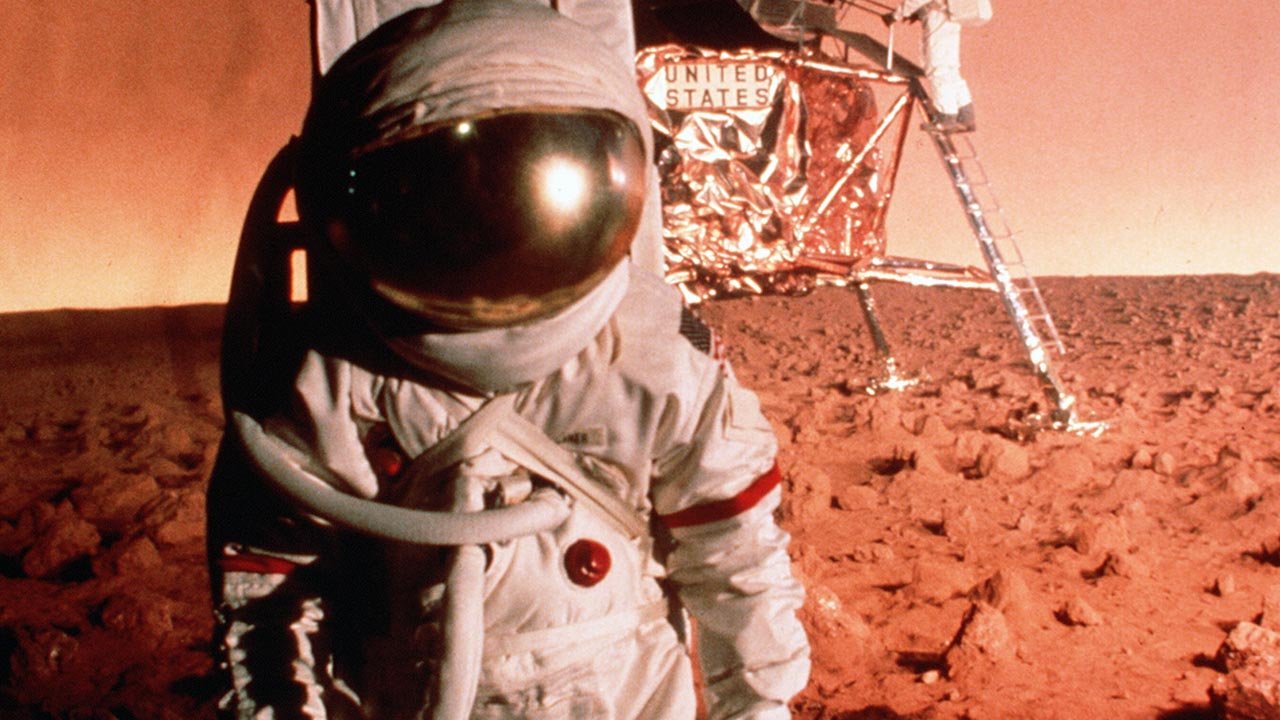 Filmstill Capricorn. Eine Person im Astronautenanzug steht auf einer rot sandigen Oberfläche. Im Hintergrund steht eine Raumkapsel.