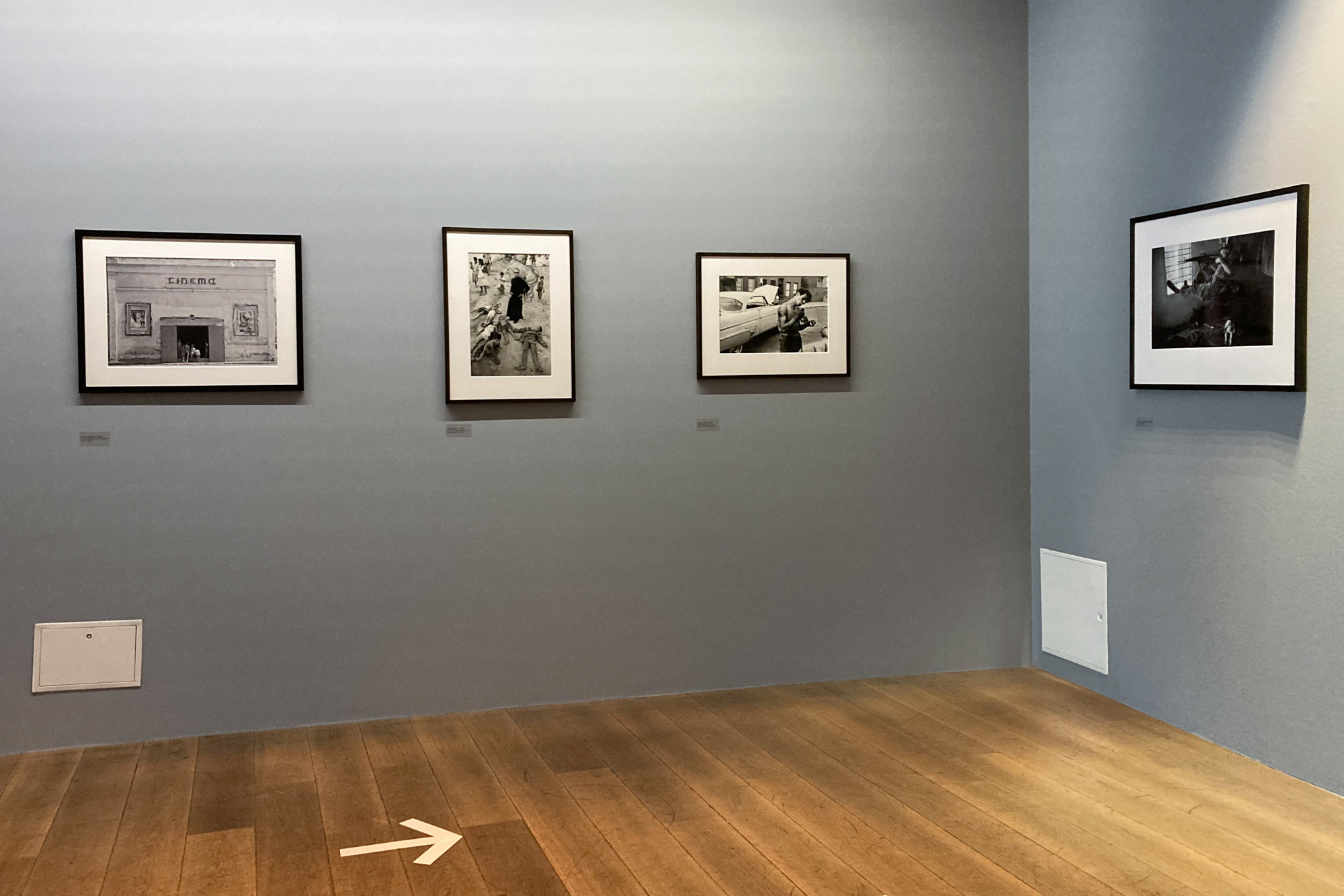 Ausstellungsfoto, welches graue Wände und Parkettboden zeigt. An den Wänden hängen schwarzgerahmte Ausstellungsstücke.
