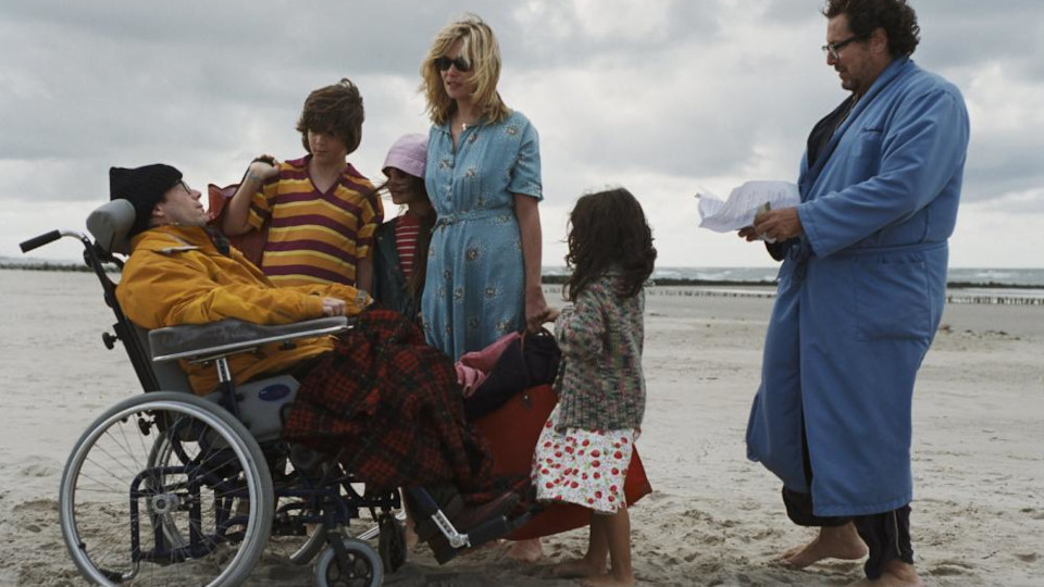 Still LE SCAPHANDRE ET LE PAPILLON Darsteller versammeln sich am Strand um eine Person im Rollstuhl