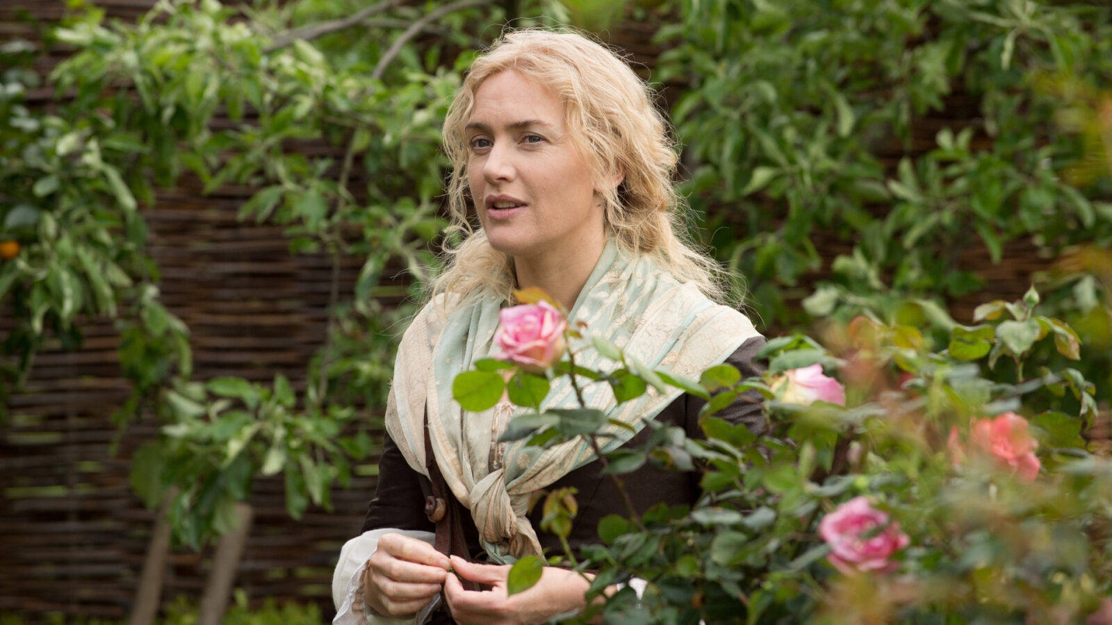 Filmstill aus Die Gärtnerin von Versailles. Eine Frau steht in mitten eines Gartens hinter einem Rosenbusch.