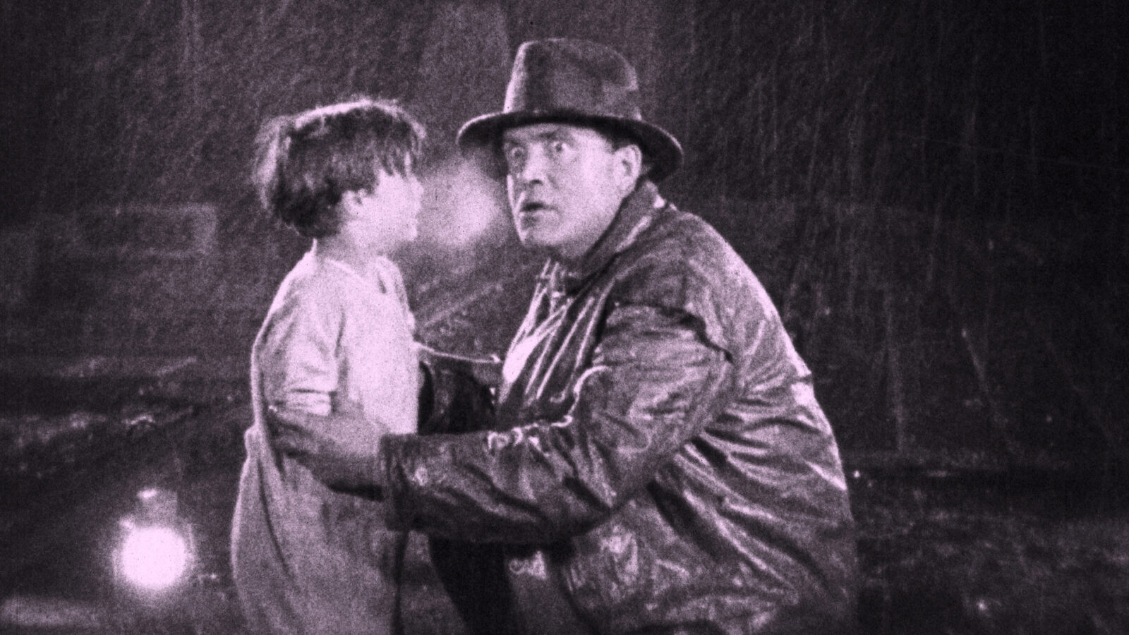 Eine Schwarz-Weiß-Aufnahme zeigt einen Mann in Lederjacke und Hut, der vor einem kleinen Jungen kniet, diesen an den Oberarmen hält.