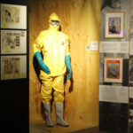 Gelber Anzug zum Schutz vor chemikalien mit blauen Handschuhen in der Ausstellung KATASTROPHE