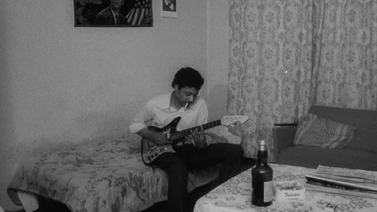 Filmstill aus They Call it Love. Auf der Schwarz-Weiß-Aufnahme sitzt ein junger Mann mit E-Gitarre auf einem Bett.