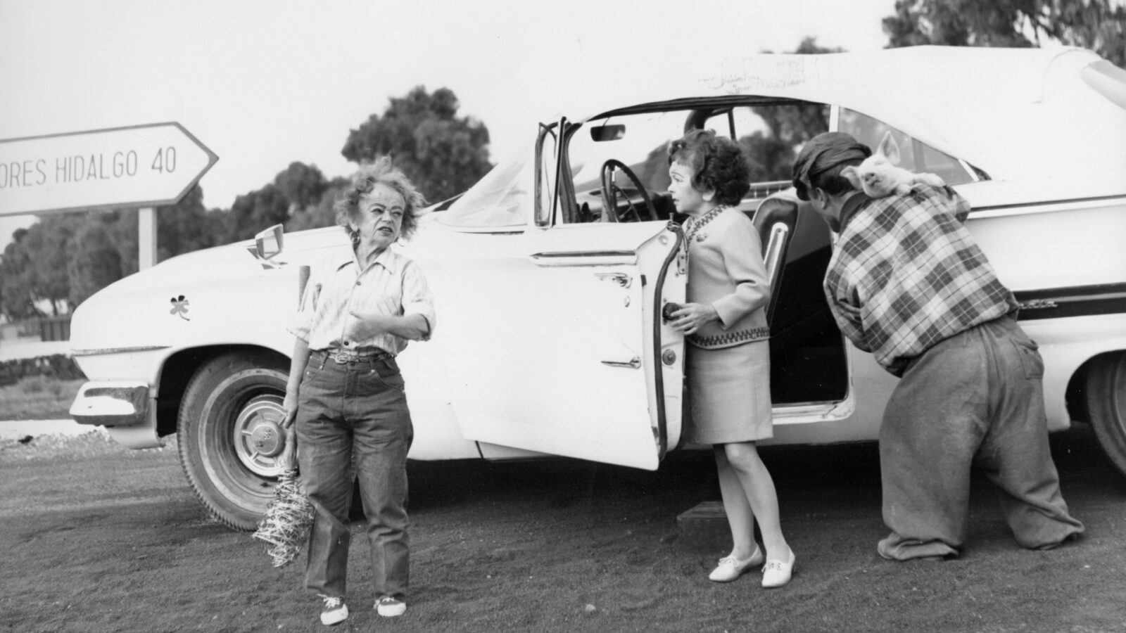 Die Schwarz-Weiß-Aufnahme zeigt drei kleinwüchsige Menschen, die vor einem weißen Auto.