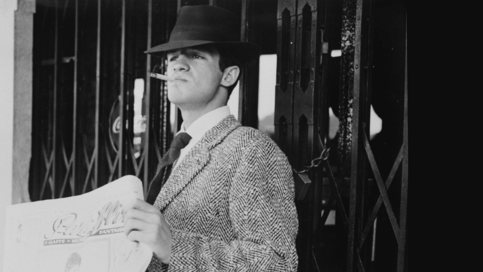 Filmstill aus dem Film Außer Atem. Ein Mann steht mit Zigarette im Mund, einer Zeitung in der Hand und Hut auf dem Kopf vor einer geschlossenen Gittertür.