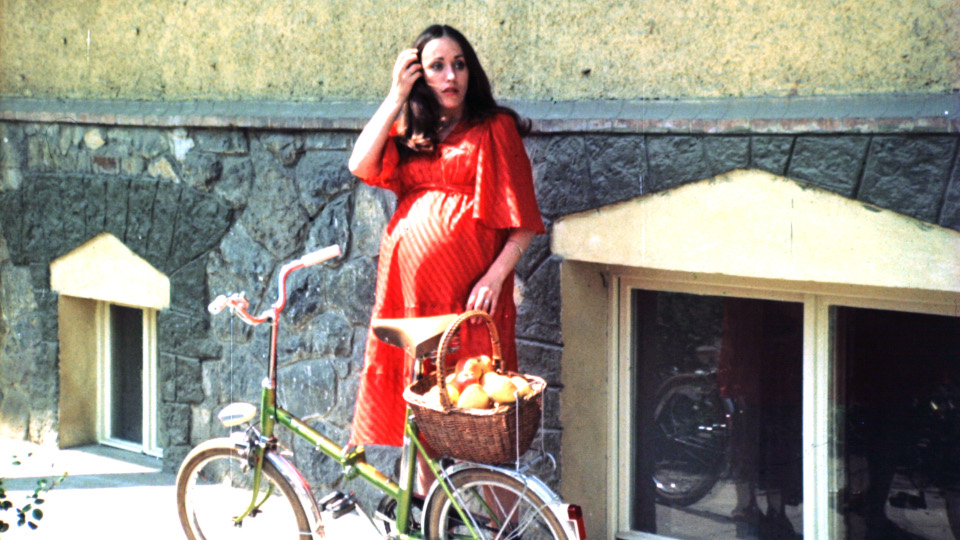 Filmstill aus Hra O Jablko. Eine schwangere Frau in einem roten Kleid steht an einer Hauswand und ein grünes Fahrrad steht vor ihr.
