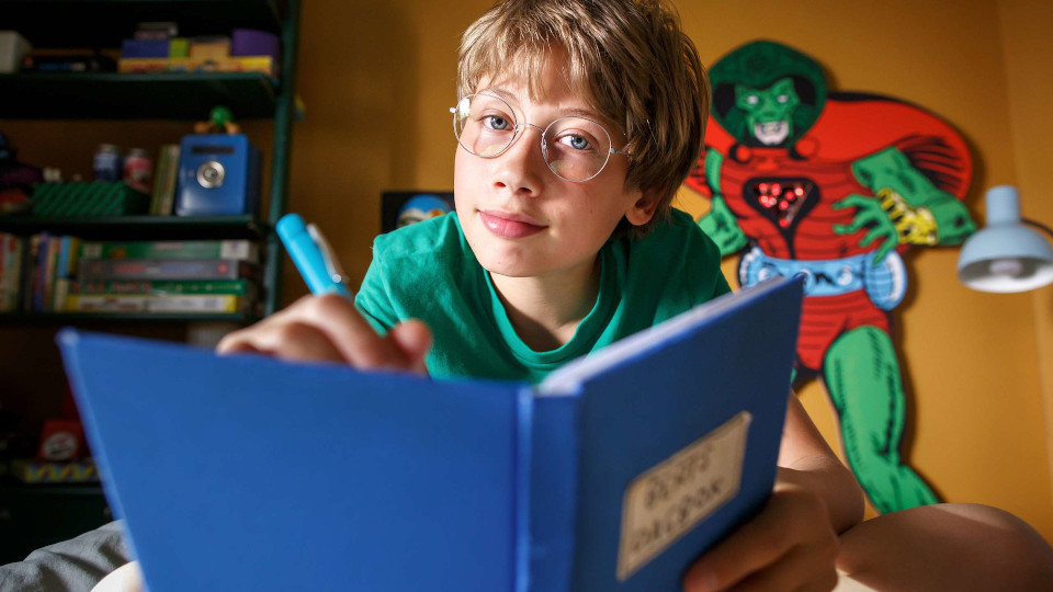 Filmstill aus Berts Katastrophen. Im Vordergrund hält eine junge ein blaues Notizbuch und einen Stift in den Händen, im Hintergrund sind ein Regal, eine Lampe und eine Superheldenfigur zu sehen.
