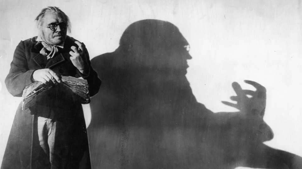 Filmstill aus Das Cabinet des Dr. Caligari. Ein älterer Mann steht vor einer Lichtquelle, die seinen gebeugten Schatten an die Wand hinter ihm wirft.