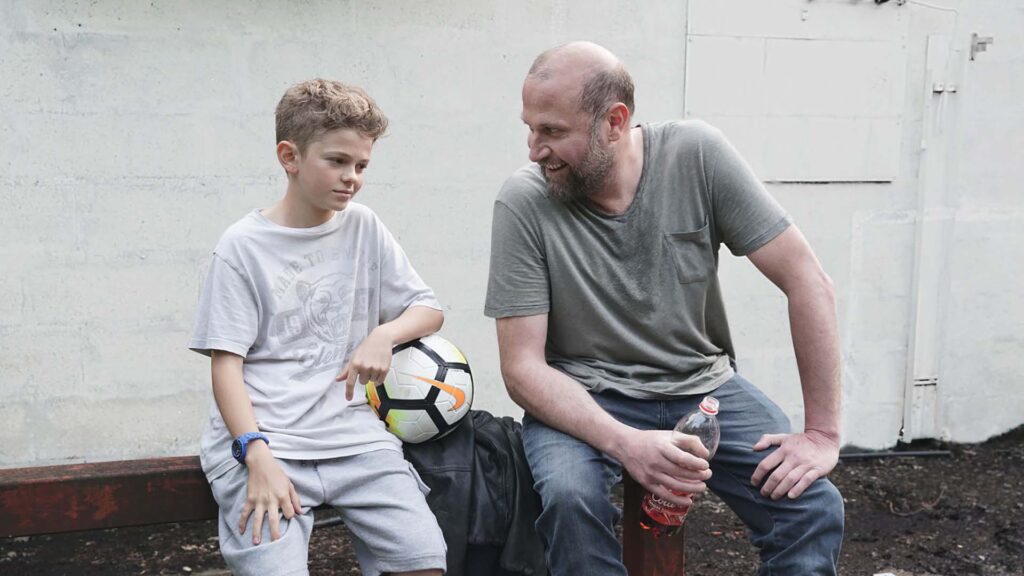 Filmstill FOURMI, Älterer Mann mit Getränk und Junge mit Fußball sitzen auf einer Mauer und unterhalten sich
