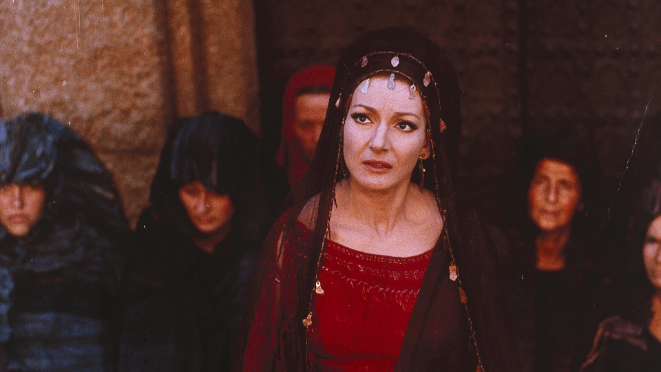 Filmstill aus Medea: Eine Frau mit Haarschmuck, im Hintergrund Frauen mit Kapuzenumhängen.