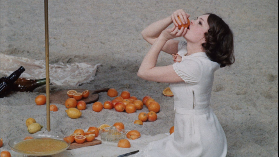 Filmstill aus Fruit of Paradise: Eine Frau in weißem Kleid sitzt im Sand und ist eine Orange mit beiden Händen
