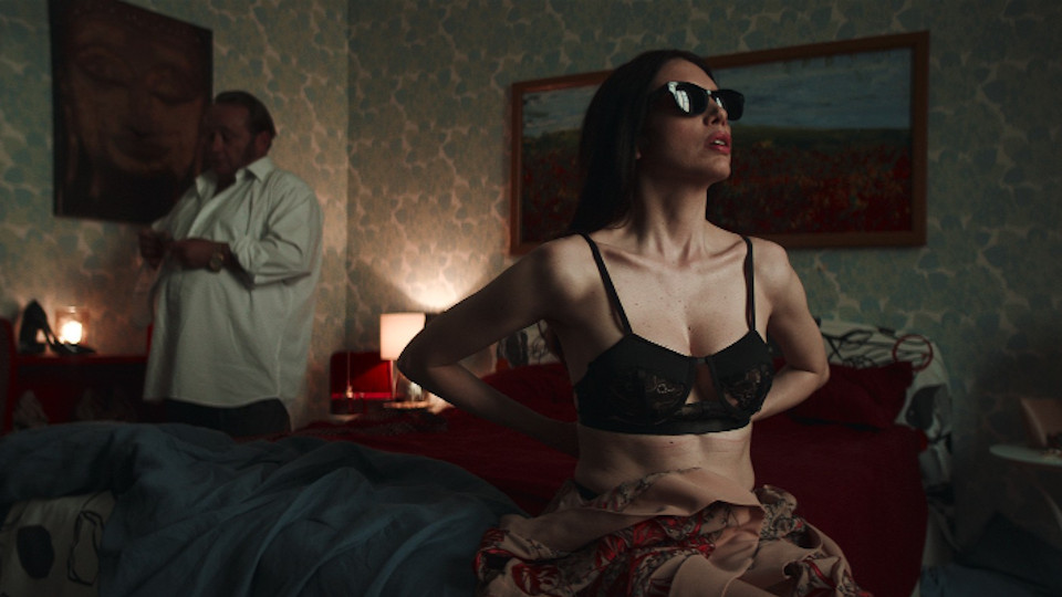 Filmstill aus Dark Glasses: Eine Frau mit Sonnenbrille sitzt in einem Bett, im Hintergrund steht ein Mann.