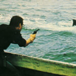 Still aus JAWS: Der Protagonist steht auf einem Boot im Meer und zielt mit einer Waffe auf die Rückenflosse des großen weißen Hais.