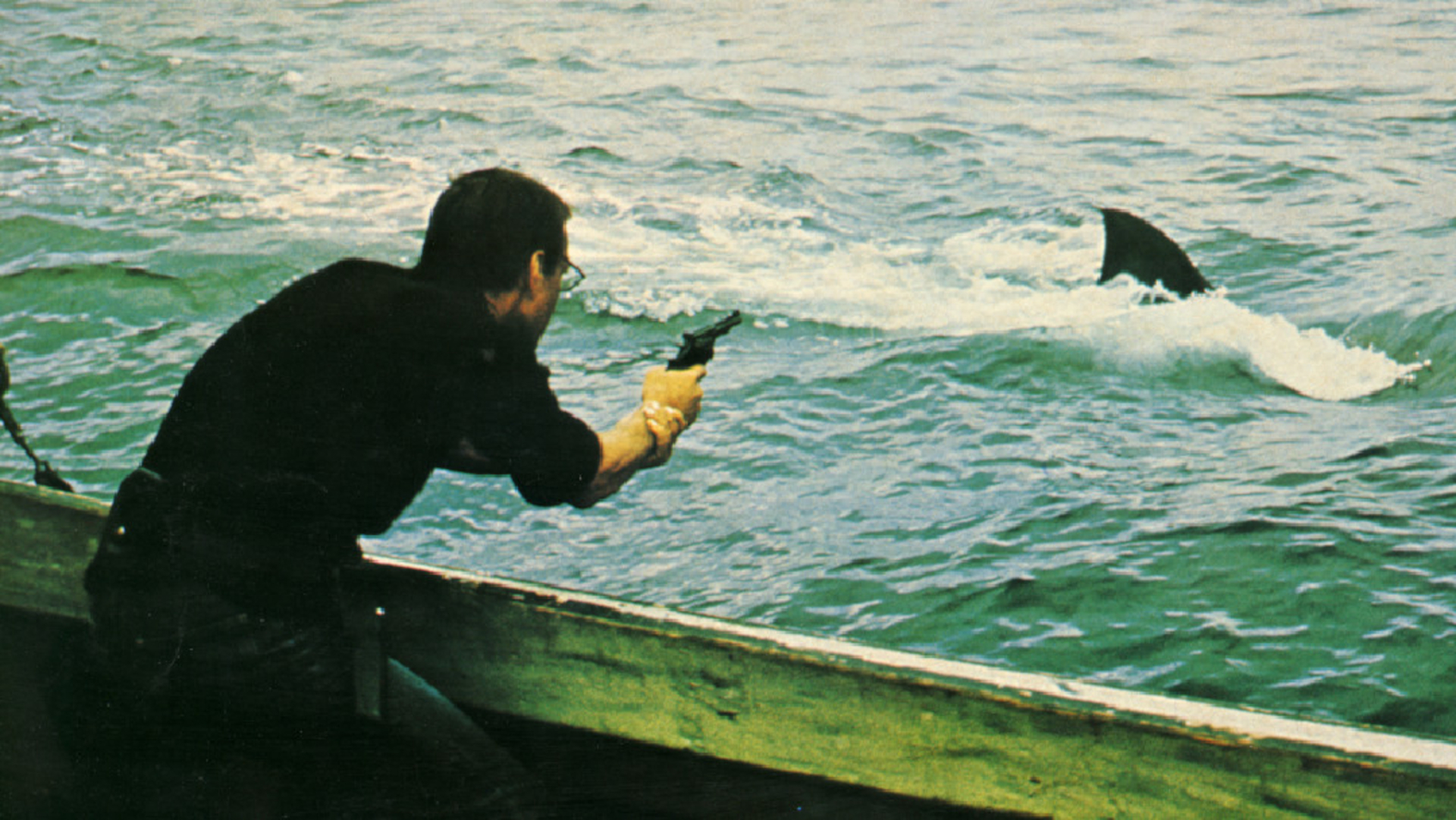 Still aus JAWS: Der Protagonist steht auf einem Boot im Meer und zielt mit einer Waffe auf die Rückenflosse des großen weißen Hais.