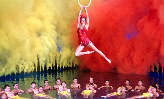 Esther Williams holding on to a ring hanging above a pool, yellow and red smoke in the background