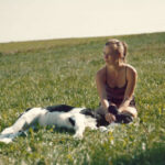 Filmstill aus NIEMAND IST BEI DEN KÄLBERN: Eine junge Frau sitzt auf einer Wiese, vor ihr liegt ein Hund
