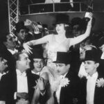Eine Frau inmitten von 15 Männern, die sie auf ihren Schultern tragen. Sie trägt ein 1920er Jahre Tanzkleid, einen Zylinder und hält eine Flasche Sekt in der Hand.