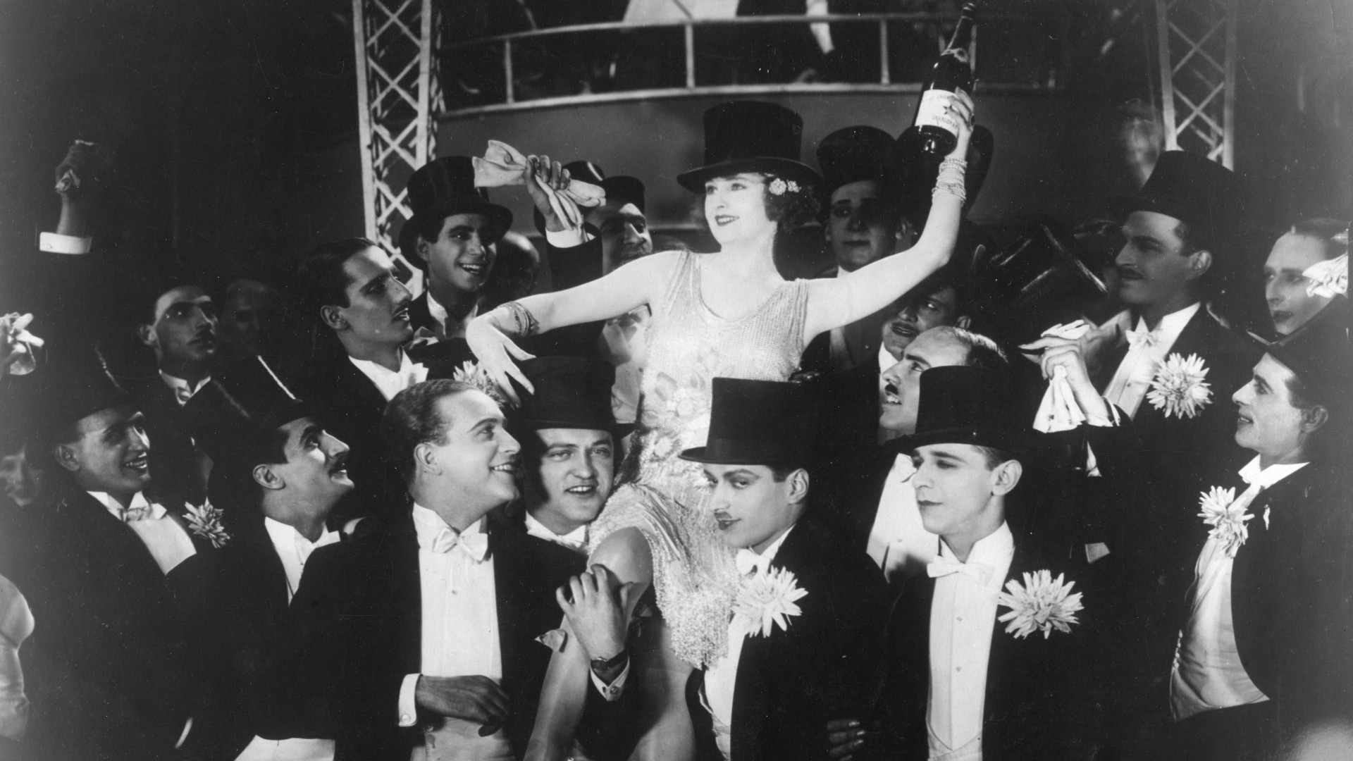 Eine Frau inmitten von 15 Männern, die sie auf ihren Schultern tragen. Sie trägt ein 1920er Jahre Tanzkleid, einen Zylinder und hält eine Flasche Sekt in der Hand.