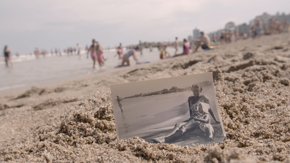 Filmstill aus Eine Frau: Ein Schwarz-Weiß-Foto einer am Strand sitzenden Frau steckt im Sand, im Hintergrund ist ein Strand zu sehen.