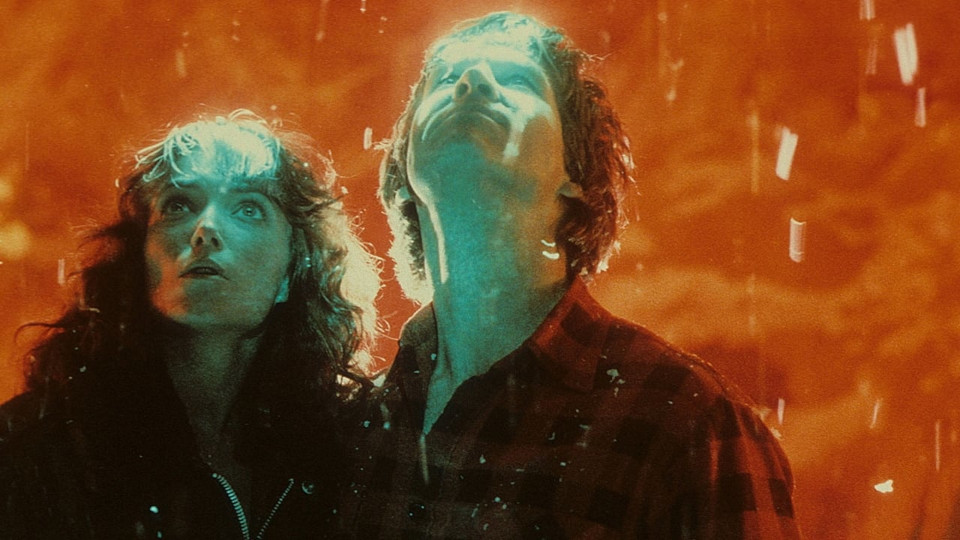 Film still from "Starman": A man and a woman in front of orange background, the man looks upwards