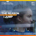 Still aus Jerry Rothwells THE REASON I JUMP: Ein junges Mädchen steht hinter einer Glasscheibe, ihr Blick ist auf etwas außerhalb des Bildes gerichtet.