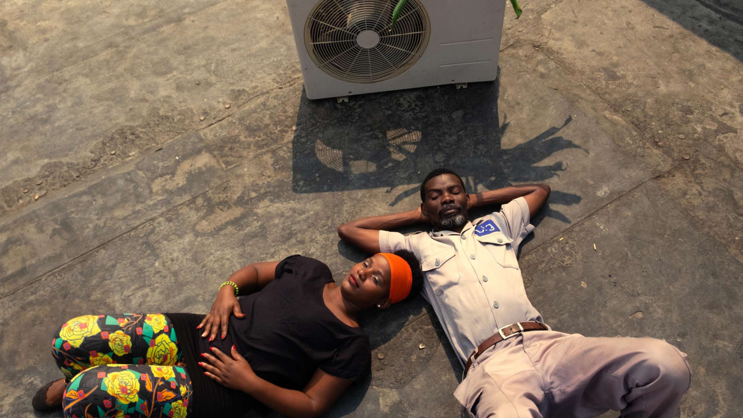 Filmstill aus Air Conditioner: Ein Mann und eine Frau liegen auf dem Boden und blicken in den Himmel, im Hintergrund steht eine Klimaanlage