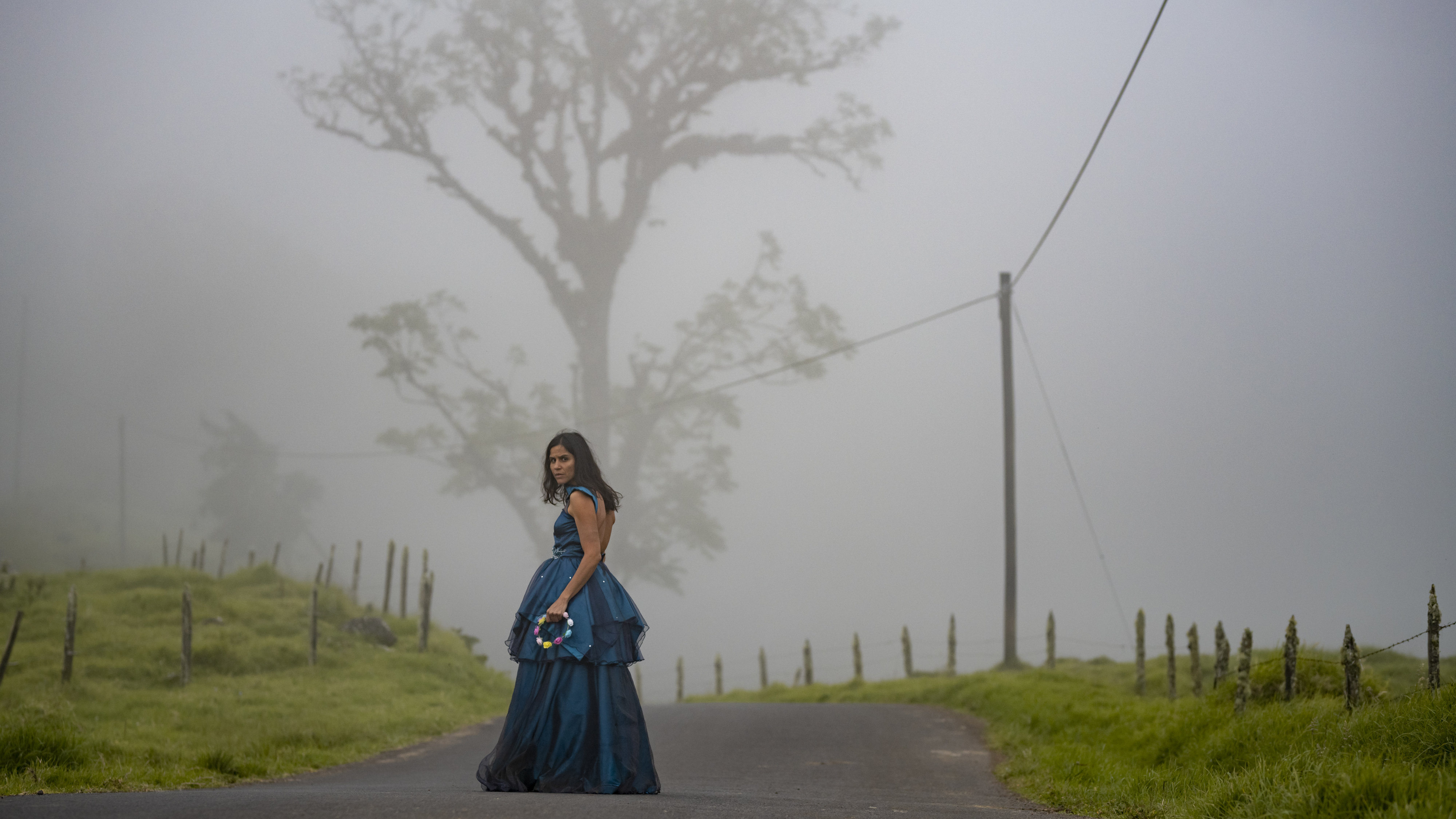 Eine junge Frau steht in einem petrolblauem Kleid auf einer Straße im Nebel und schaut über ihre Schulter zurück.