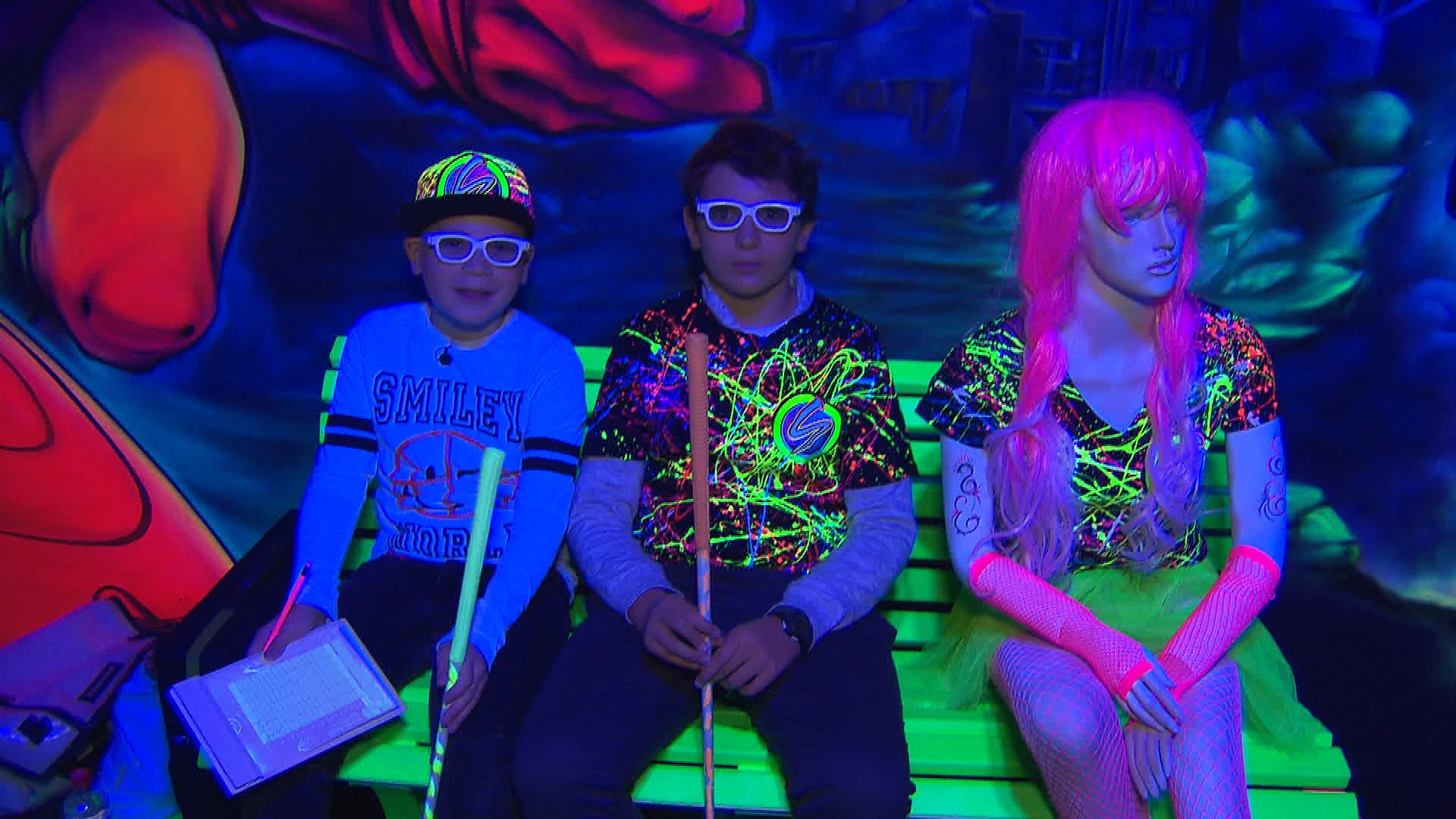 Drei Personen sitzen in einer abgedunkelten Umgebung, in der einzelne neonfarbene Elemente hervorstechen.