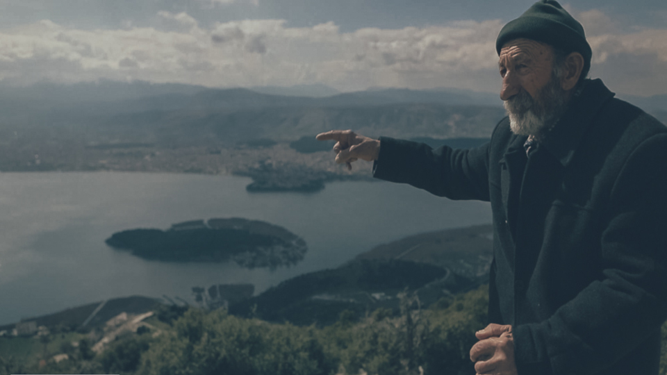 Filmstill aus TO BALKONI: Ein Mann zeigt mit seinem rechten Arm auf etwas in der Ferne, im Hintergrund sieht man eine Landschaft mit einem See.