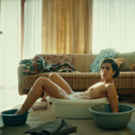 Filmstill aus Wir könnten genauso gut tot sein: Eine Frau sitzt in einem Wohnzimmer nackt in einer kleinen Wanne mit Badewasser, ihre Füße und Arme sind in kleinen Schüsseln mit Badewasser
