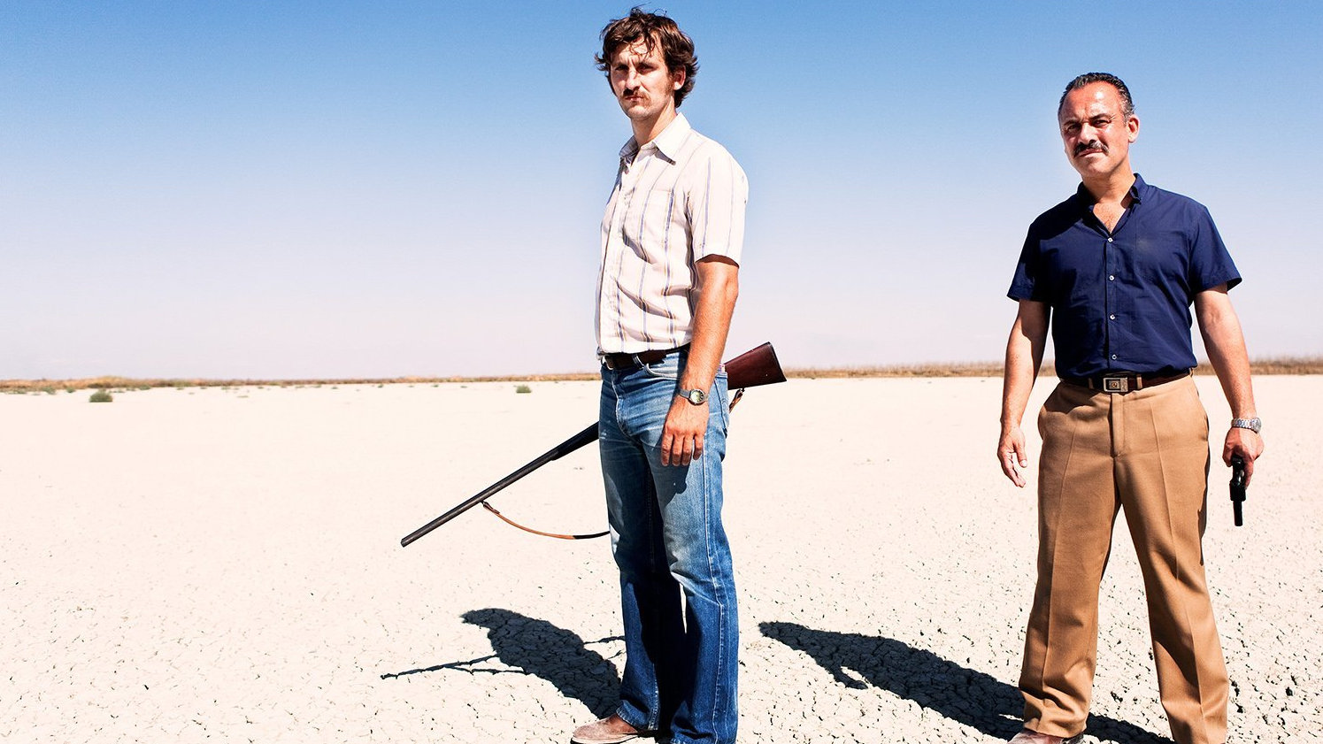 Ein Filmstill zeigt zwei Männer mit Pistole und Gewehr in der Wüste.