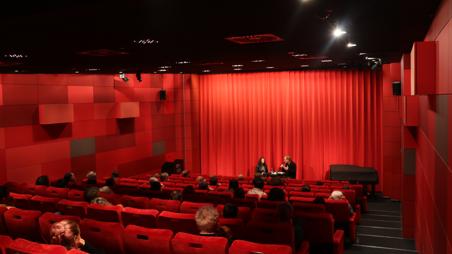 Das Kino des DFF: Rote Samtstühle, rote Wand, roter Vorhang; in den Reihen sitzen Zuschauer:innen und vor dem geschlossenen Vorhang sitzen zwei Personen, die in einem Gespräch sind