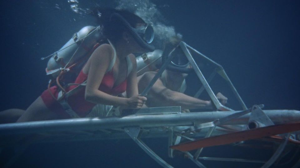 Filmstill aus Underwater! Eine Frau hält eine Metallkonstruktion vor sich, während sie mir Taucherbrille, -maske und Sauerstoffflasche hinter her schwimmt.