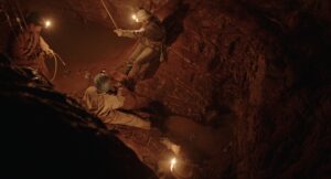 Filmstill aus IL BUCO. Drei Personen sind in Arbeitsanzügen und mit Stirnlampen in einer Höhle zu sehen. 