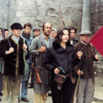 Filmstil, das eine Gruppe Menschen zeigt, die bewaffnet sind und eine rot schwarze Fahne vor sich her tragen.